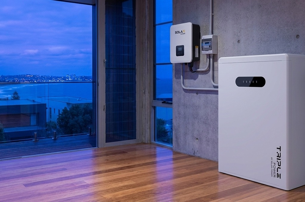 Close crop of a SolaX hybrid inverter and Triple Power battery mounted on a wall with an ocean   view through the window behind, ready to join a virtual power plant in Australia