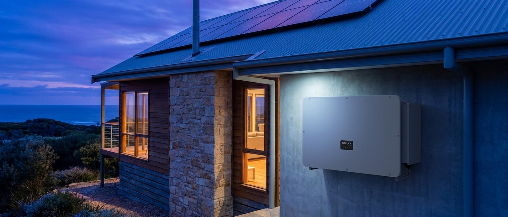 SolaX Power inverter mounted on the exterior render wall of a coastal stone home at dusk with solar panels on the roof.