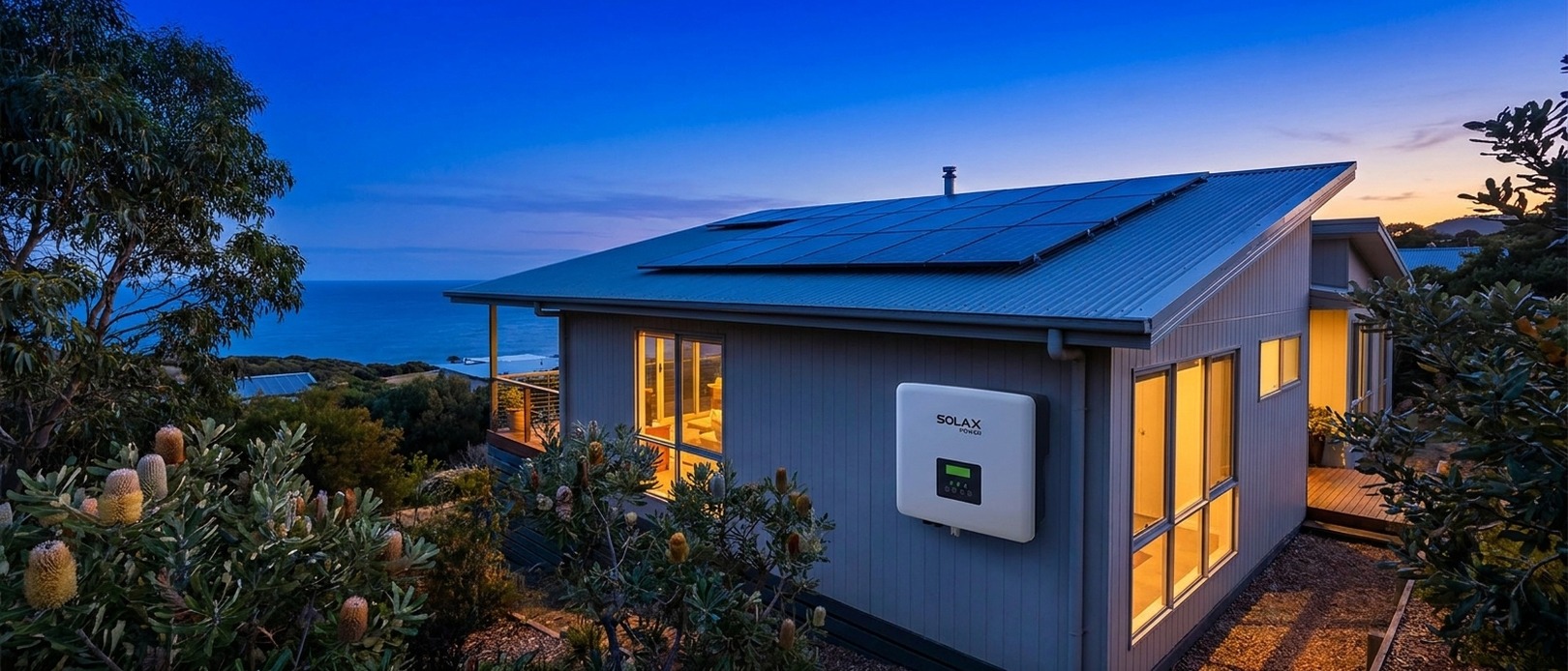 SolaX Power inverter on the exterior wall of a coastal home at dusk with solar panels on a metal roof and native banksia shrubs in the foreground.