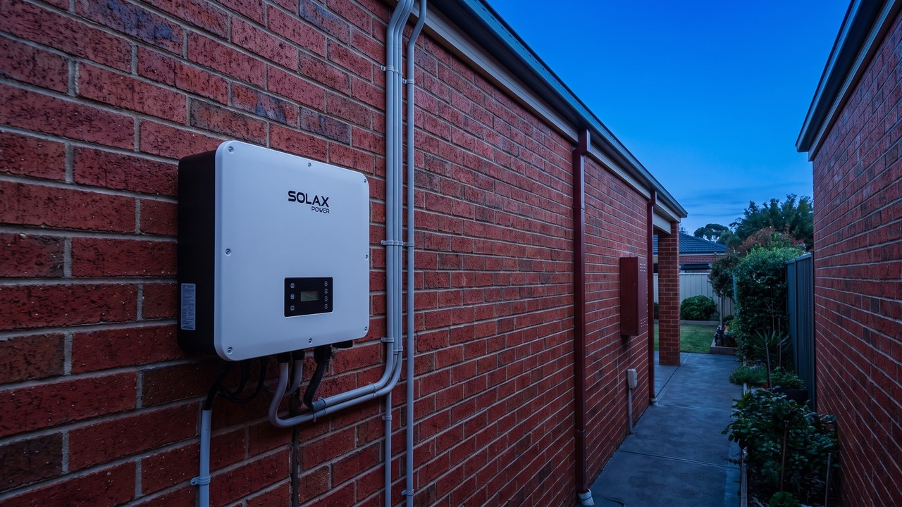 SolaX inverter wall-mounted on a red brick exterior wall of an Australian home at dusk, with a path and garden leading down the side of the house
